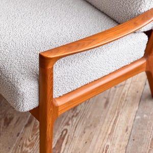 Puede incluir: Primer plano de un sill&oacute;n moderno de mediados de siglo. La silla presenta tapicer&iacute;a texturizada de color gris claro y reposabrazos y patas de madera de tonos c&aacute;lidos. El marco de madera tiene un dise&ntilde;o suave y curvado.