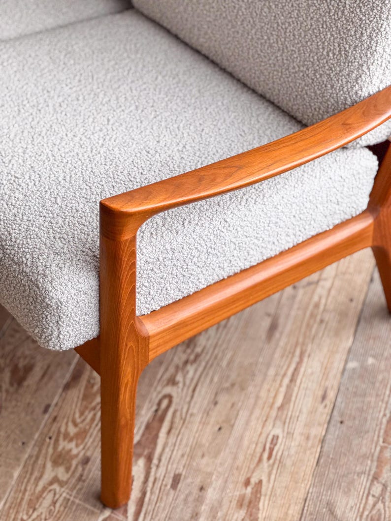 Puede incluir: Primer plano de un sill&oacute;n moderno de mediados de siglo. La silla presenta tapicer&iacute;a texturizada de color gris claro y reposabrazos y patas de madera de tonos c&aacute;lidos. El marco de madera tiene un dise&ntilde;o suave y curvado.