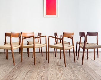 May include: A set of seven mid-century modern chairs with woven seats and wooden frames. The chairs have a curved backrest and armrests. The chairs are arranged on a wooden floor in front of a white wall with a framed artwork.