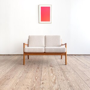 Puede incluir: Un sof&aacute; de dos plazas de estilo moderno de mediados de siglo, con una tela clara y texturizada y un marco de madera. El sof&aacute; est&aacute; colocado contra una pared blanca, con una obra de arte enmarcada que muestra un degradado de tonos rosas y rojos. El suelo es de tablones de madera.