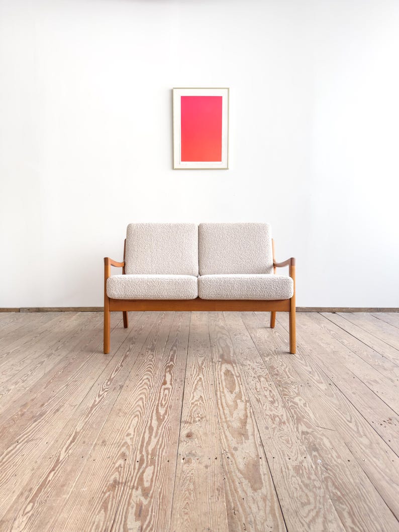 Puede incluir: Un sof&aacute; de dos plazas de estilo moderno de mediados de siglo, con una tela clara y texturizada y un marco de madera. El sof&aacute; est&aacute; colocado contra una pared blanca, con una obra de arte enmarcada que muestra un degradado de tonos rosas y rojos. El suelo es de tablones de madera.