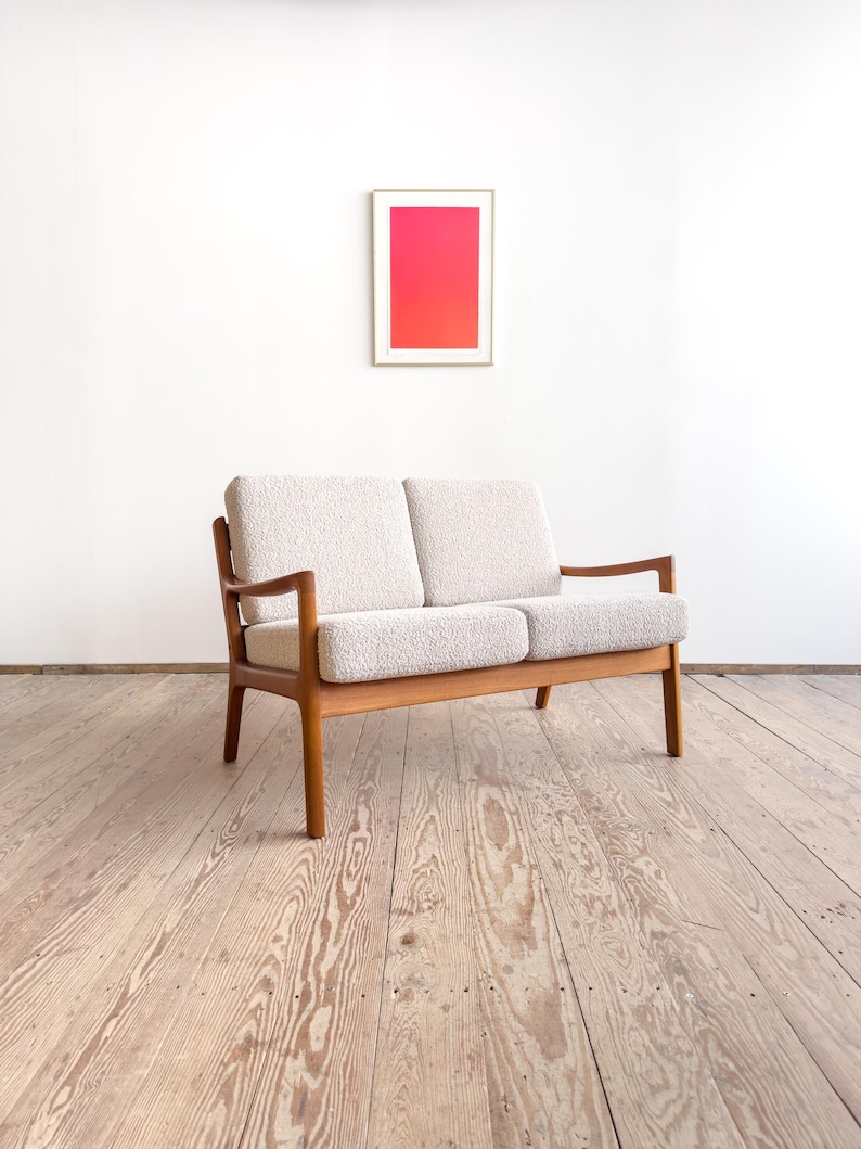 Puede incluir: Un sof&aacute; de dos plazas de estilo moderno de mediados de siglo, con una tela clara y texturizada y un marco de madera. El sof&aacute; tiene un dise&ntilde;o minimalista. Una obra de arte enmarcada con un degradado de rosa y rojo cuelga de la pared blanca detr&aacute;s del sof&aacute;.