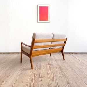 Puede incluir: Un sof&aacute; de dos plazas de estilo moderno de mediados de siglo, con estructura de madera clara y tapicer&iacute;a de color claro. El sof&aacute; tiene respaldo de listones y patas anguladas. Una obra de arte enmarcada con un degradado rosa y rojo cuelga de la pared blanca detr&aacute;s del sof&aacute;.