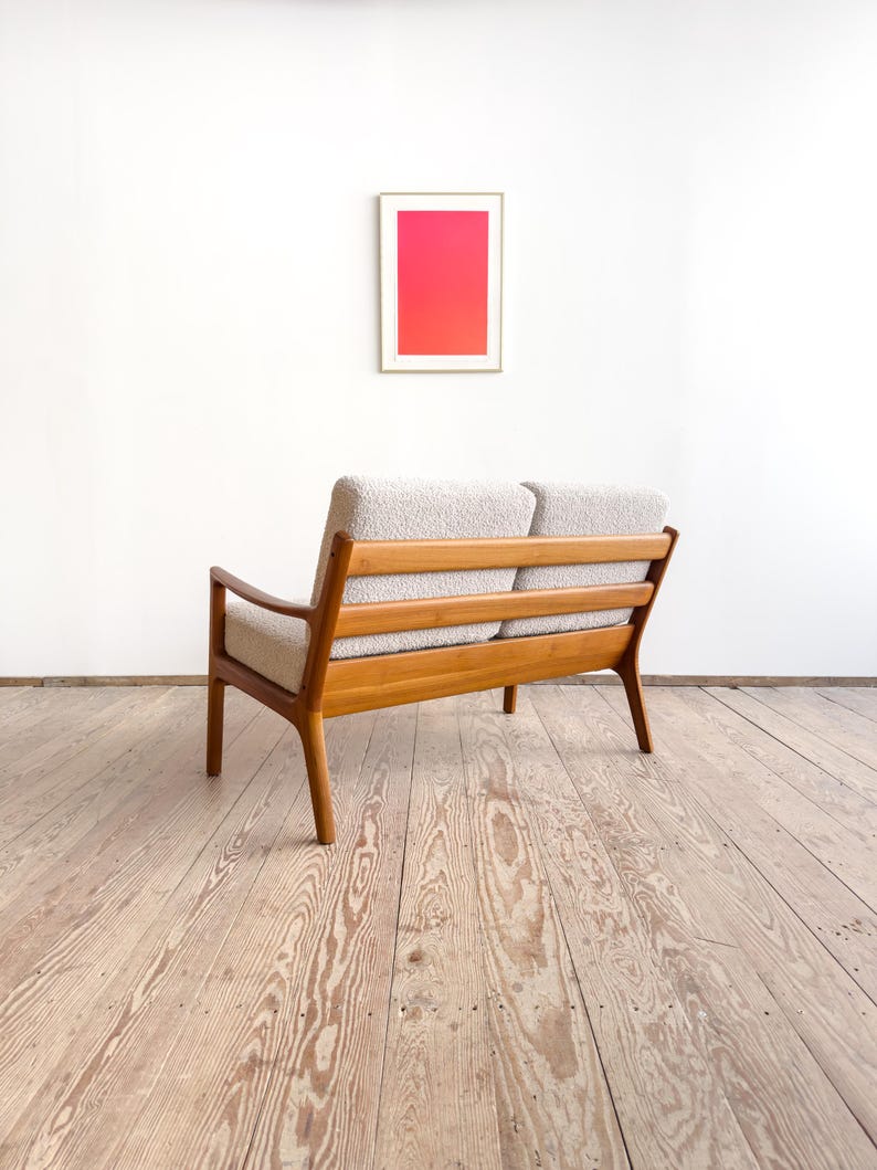 Puede incluir: Un sof&aacute; de dos plazas de estilo moderno de mediados de siglo, con estructura de madera clara y tapicer&iacute;a de color claro. El sof&aacute; tiene respaldo de listones y patas anguladas. Una obra de arte enmarcada con un degradado rosa y rojo cuelga de la pared blanca detr&aacute;s del sof&aacute;.