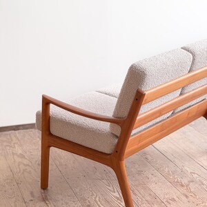 Puede incluir: Un sof&aacute; de estilo moderno de mediados de siglo con asiento y respaldo de tela clara y texturizada. El sof&aacute; tiene una estructura de madera con acabado natural y reposabrazos curvos. El dise&ntilde;o es minimalista.