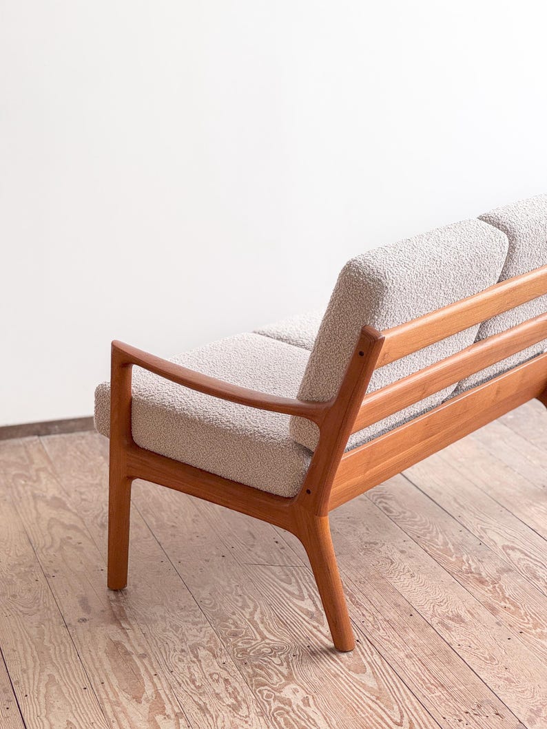 Puede incluir: Un sof&aacute; de estilo moderno de mediados de siglo con asiento y respaldo de tela clara y texturizada. El sof&aacute; tiene una estructura de madera con acabado natural y reposabrazos curvos. El dise&ntilde;o es minimalista.