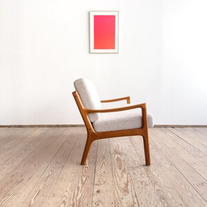 Puede incluir: Un sill&oacute;n retro con asiento texturizado de color claro y estructura de madera. La silla est&aacute; colocada frente a una pared blanca con una obra de arte enmarcada que presenta un degradado de tonos rosas y rojos. El dise&ntilde;o de la silla es de mediados del siglo XX.