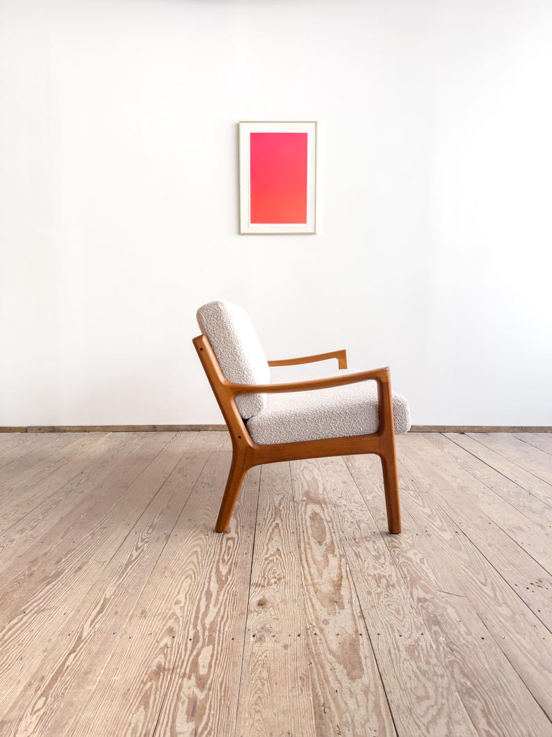 Puede incluir: Un sill&oacute;n retro con asiento texturizado de color claro y estructura de madera. La silla est&aacute; colocada frente a una pared blanca con una obra de arte enmarcada que presenta un degradado de tonos rosas y rojos. El dise&ntilde;o de la silla es de mediados del siglo XX.