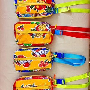 May include: Six rectangular waist bags featuring a vibrant yellow background with a colorful fruit and floral pattern. Each bag includes a zippered main compartment and an adjustable nylon strap with a plastic buckle in lime green, red, or blue.
