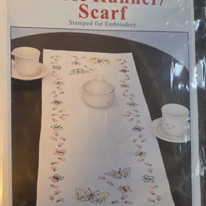 May include: White table runner/scarf stamped for embroidery, featuring a butterfly design. The runner measures 15" x 42" and is made of cotton/poly broadcloth. Includes a small white bowl and two teacups. The text on the packaging reads "Table Runner/Scarf".