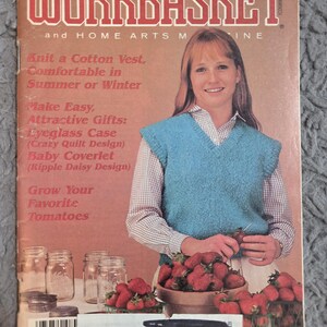 May include: The Workbasket magazine cover from May 1986. The cover features a woman wearing a blue knit vest. Text includes "Knit a Cotton Vest" and "Grow Your Favorite Tomatoes". Several baskets of strawberries are in the foreground.
