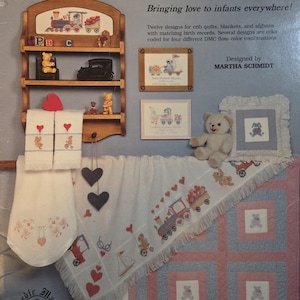 May include: A promotional image for baby-themed needlepoint patterns. The image features a wooden shelf with embroidered designs, including a train and teddy bears. A quilt with teddy bear squares and a matching blanket with fringe are also shown. The text reads "Covers for Cuddlin".