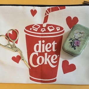 May include: A white zippered pouch with a red "diet Coke" design and red hearts. A gold-colored scissor and a small green floral-patterned tin are on the pouch.