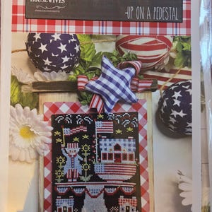 May include: A cross-stitch pattern titled "Americana Bakery - Up on a Pedestal" featuring patriotic imagery. The design includes a red, white, and blue color scheme with stars, flags, and a bakery theme. The background is a red and white checkered pattern.