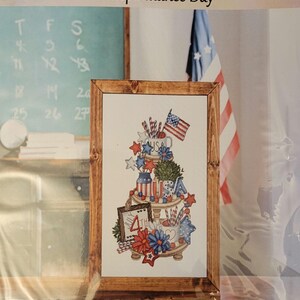 May include: Cross-stitch pattern for Independence Day. The design features a tiered display with patriotic elements like the American flag, stars, and red, white, and blue decorations. The framed artwork is labeled "Seasonal Collection" and "Independence Day".