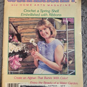 May include: The Workbasket and Home Arts Magazine cover from April 1984. The cover features a woman wearing a blue crochet top, holding a flower. The magazine's title is in blue and pink. The cover also includes text about crochet and creating an afghan.