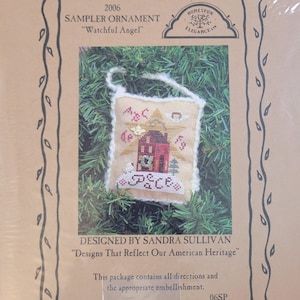 May include: A sampler ornament kit titled "Watchful Angel" featuring a cross-stitch design of a red house, a star, and the word "Peace". The kit includes directions and embellishments. The design is by Sandra Sullivan.