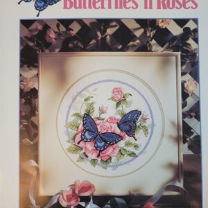 May include: A cross-stitch pattern titled "Butterflies 'n Roses" by Paul Sweany. The design features two blue butterflies and pink roses within a circular frame. The Dimensions logo is at the bottom.