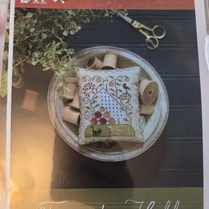 May include: A cross-stitch sampler kit with the text "Plum Street Samplers" and "Tomato Hill." The kit includes a small, embroidered pillow with a tomato and bird design, surrounded by wooden spools and scissors.