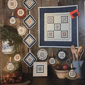 May include: A collection of country patchwork items, including quilted squares, pot holders, and decorative brooms, arranged on a wooden surface. The items feature floral embroidery in red, blue, and white. The image includes the text "Country Patchwork" and "BOOK 20".