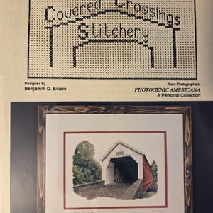 May include: A framed print of a covered bridge with a red roof and white siding, set against a backdrop of green trees. The frame is a dark wood. Above the print is a cross-stitch pattern with the words "Covered Crossings Stitchery".