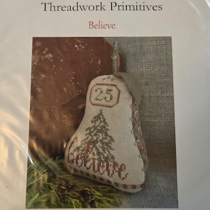 May include: A pear-shaped fabric ornament featuring cross-stitch embroidery of a Christmas tree, the number 25, and the word Believe. The design includes a stem and leaf, with decorative stitching along the side edges and a striped base.