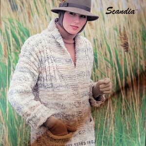May include: A beige knitted cardigan with a cable knit pattern and a button closure. The cardigan has a large pocket on the front and is worn by a woman with a brown hat and gloves. The text "Scandia" is visible in the top right corner of the image.