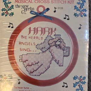 May include: A musical cross stitch kit featuring a pink and white design of an angel holding a musical instrument. The text reads "Hark the Herald Angels Sing." The kit includes a musical chip.