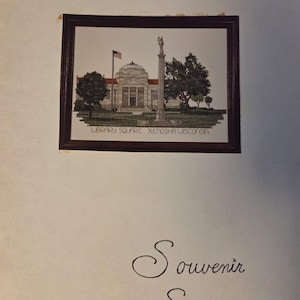 May include: A framed embroidery of the Library Square in Kenosha, Wisconsin, featuring a domed building, an American flag, and a monument. The embroidery is in a dark brown frame. The words "LIBRARY SQUARE KENOSHA WISCONSIN" are at the bottom of the image. The words "Souvenir Stitchery" are below the frame.