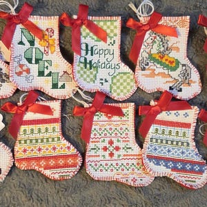 May include: A collection of small, cross-stitched Christmas stockings with red ribbon bows. Some stockings feature the words "NOEL" and "Happy Holidays", while others display festive patterns and images of birds, a horse, and a dog.