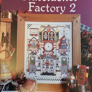 May include: A framed cross-stitch pattern titled "The Nutcracker Factory 2" with a Christmas theme. The pattern features a detailed clock tower design with nutcracker figures, houses, and the text "Merry Christmas".