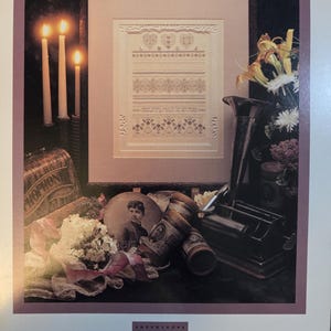 May include: Framed cross-stitch sampler with a floral and heart design. The sampler is in a dark wooden frame. The image includes lit candles, flowers, and vintage items. The text "Shepherd's Bush Printworks" is visible.