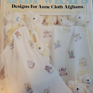 May include: A soft, white baby blanket with embroidered designs of teddy bears and other baby-themed images. The blanket is draped over a crib with white teddy bears and a pacifier. The text "BABY WRAPS" and "Designs For Anne Cloth Afghans" is visible.