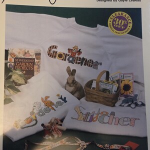 May include: A white sweatshirt with the word "Gardener" embroidered in a patchwork style, featuring a sunflower and a scarecrow. Another sweatshirt says "Stitcher". The image also includes a rabbit, a basket, and gardening-themed items.