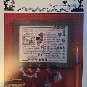 May include: A cross-stitch sampler titled "Cross-Stitcher's Sampler" by Sigrid Designs, framed and displayed on a red wall. The sampler features text and images related to cross-stitching, with a candle and decorative items in the foreground.