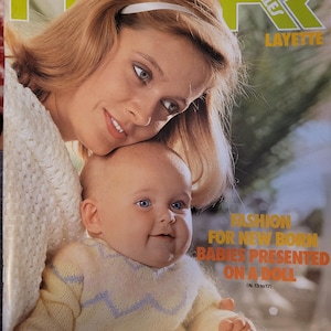 May include: Magazine cover featuring a woman and a baby. The baby wears a yellow sweater with a blue zigzag pattern. The magazine title is "PHILDAR MAILLES LAYETTE" with the text "FASHION FOR NEW BORN BABIES PRESENTED ON A DOLL".