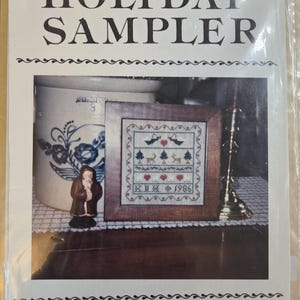 May include: The image shows a framed cross-stitch sampler with a holiday theme. The sampler features birds, trees, hearts, and the year 1986. The frame is wooden, and the background includes a ceramic jug and a small figurine. The text "THE HOLIDAY SAMPLER" is at the top.