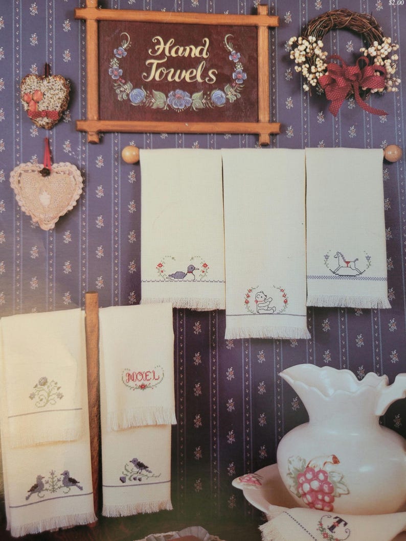 May include: A set of six white hand towels with embroidered designs. The designs include a bird, a teddy bear, a rocking horse, a floral design, the word "NOEL", and a bird with a sprig of holly. The towels are hanging on a rack against a purple and white patterned wallpaper.