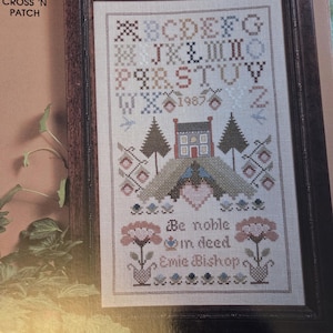 May include: A framed cross-stitch sampler with the text "NOBLE DEED" at the top. The sampler features an alphabet, a house, trees, and the words "Be noble in deed, Emie Bishop". The frame is brown.