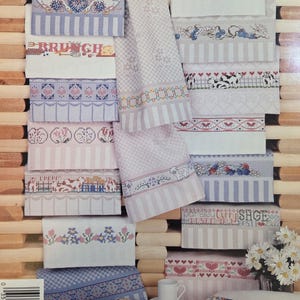 May include: Embroidered kitchen towels in a variety of designs, including floral patterns and the word "BRUNCH". The towels are in shades of pink, blue, and white, with a wooden background. The towels are stacked.