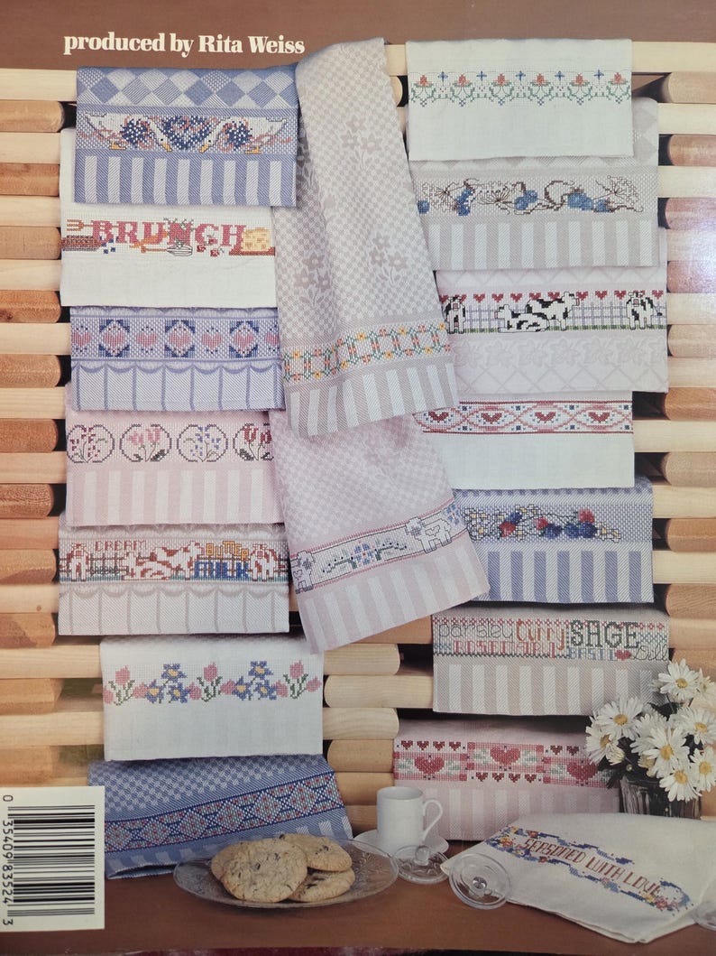 May include: Embroidered kitchen towels in a variety of designs, including floral patterns and the word "BRUNCH". The towels are in shades of pink, blue, and white, with a wooden background. The towels are stacked.