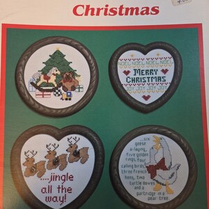 May include: A Christmas-themed cross-stitch pattern booklet titled "A Merry Keeper Christmas." The cover features four framed cross-stitch designs: a Christmas tree, a heart with "Merry Christmas," reindeer, and a goose with text.