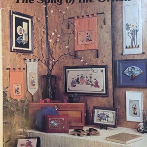 May include: An advertisement for needlepoint and cross stitch designs. The image features framed artwork, banners, and decorative items with an Asian theme. The text reads "This is 'The Song of the Orient'" and includes the designer's name and contact information.