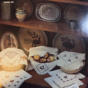 May include: A display of vintage-style cross-stitch patterns and finished pieces. The image features a variety of embroidered linens, including napkins and basket liners, alongside decorative plates and a serving spoon. The text "Petite Fleurs" is visible.