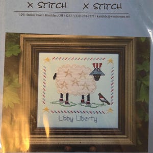 May include: Cross-stitch pattern featuring a sheep with star accents, wearing a patriotic hat, and the text "Libby Liberty." The design includes a bird and is framed in a wooden border. The brand name "Katidid" is at the top.