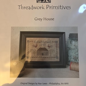 May include: A framed cross-stitch sampler titled "Grey House" by Threadwork Primitives. The sampler features a house, roosters, and the text "Established 1799." The frame is dark, and the background is a neutral color. A jar and thread spool are visible.