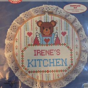 May include: A counted cross stitch kit featuring a teddy bear design with the text "IRENE'S KITCHEN." The kit includes a wooden hoop, a white lace trim, and the Colortex logo. Finished size is 10.25 inches.