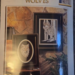 May include: A package featuring framed cross-stitch art with timber wolf designs. The artwork includes a framed oval portrait of a wolf's head and a framed image of a wolf in a forest setting. The package has the text "TIMBER WOLVES".