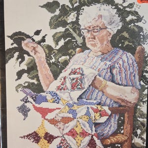 May include: A cross-stitch pattern kit titled Silver Threads & Golden Memories featuring an illustration of an older person with white hair and glasses sitting in a wooden chair, sewing a colorful quilt with red, blue, and yellow geometric patterns.