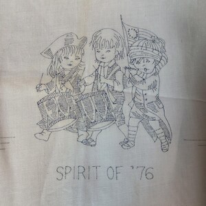 May include: Off-white fabric with a blue embroidered design. The design features three children marching, playing drums and a fife, and carrying a flag. The text "SPIRIT OF '76" is embroidered below the figures. The approximate size is 16 x 42 inches.